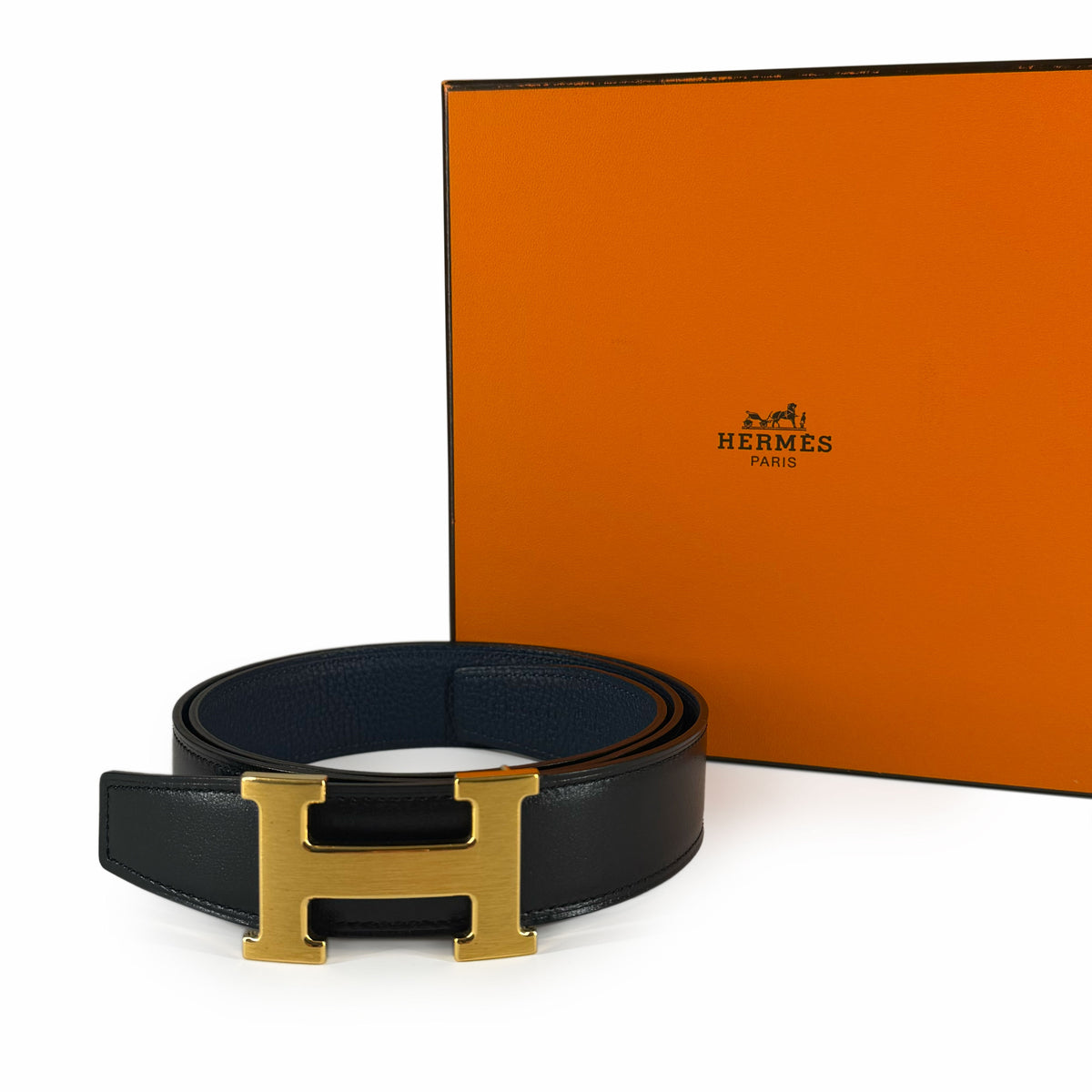 Black leather Hermès belt with gold-tone H buckle and orange brand box, curated Pre-loved Luxury by Myluxe in Helsinki.
