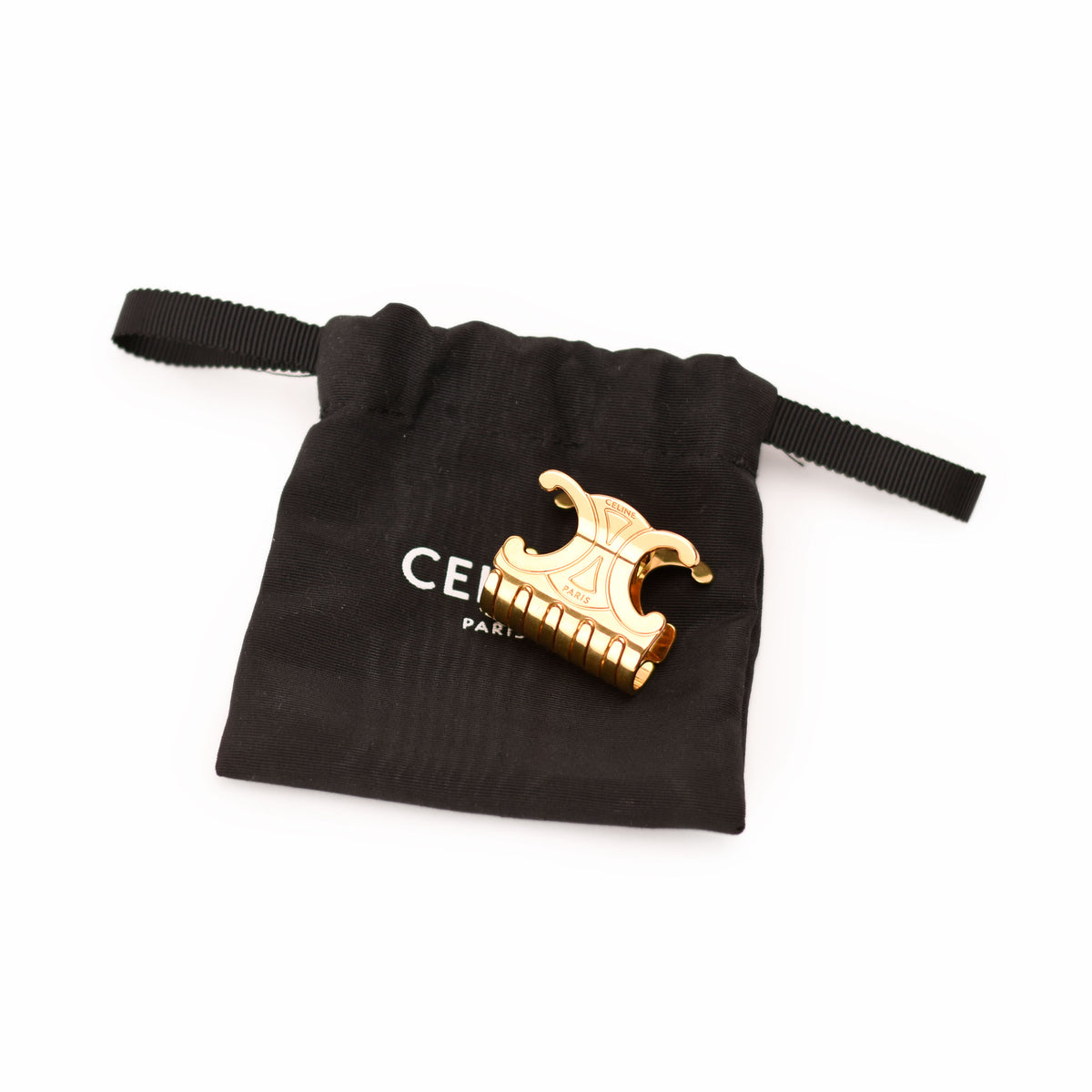 A gold-tone metal hair claw clip with an ornate scroll design, displayed on top of a black drawstring pouch with white Celine branding, set against a plain white background.