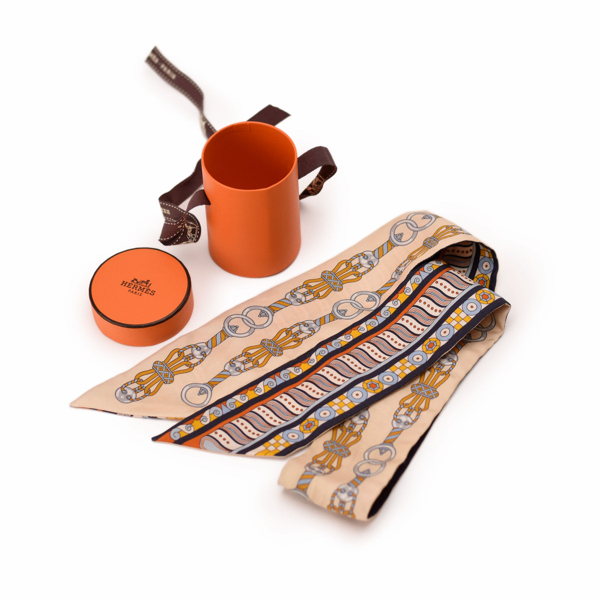 Hermès silk scarf with orange, blue, and gold geometric patterns and matching orange gift box. curated Pre-loved Luxury by Myluxe in Helsinki.